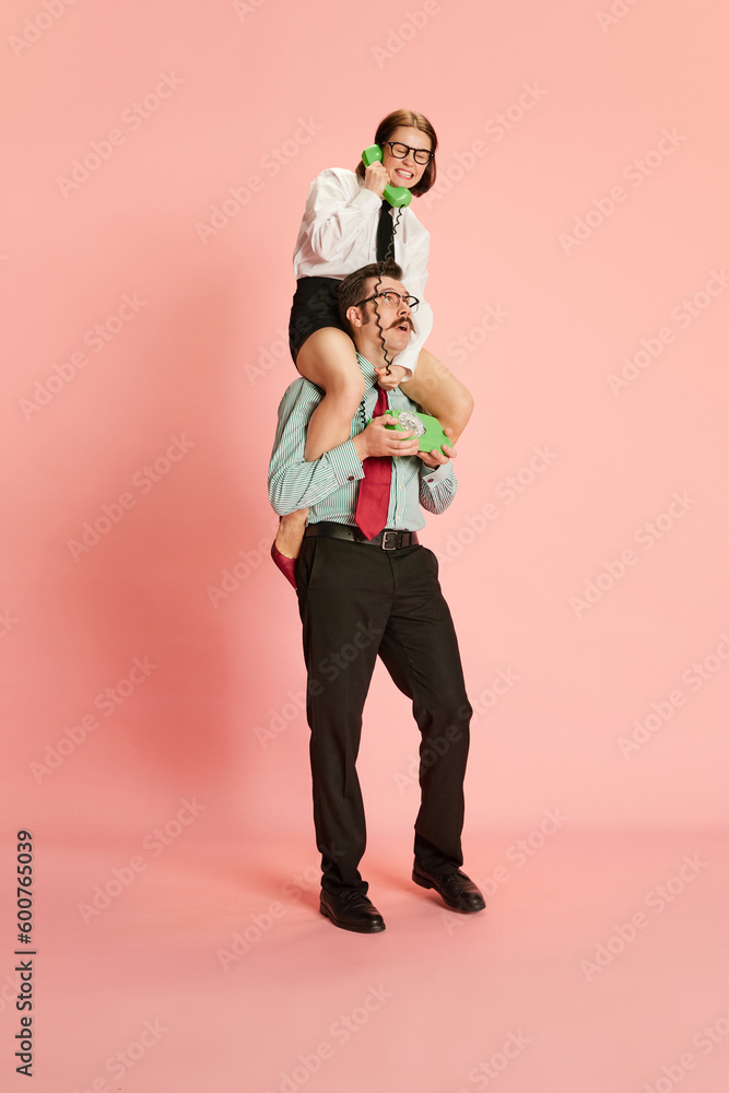 Obraz premium Portrait with couple, shocked man and emotional woman sitting on male shoulders speaking via telephone and shouting over pink studio background