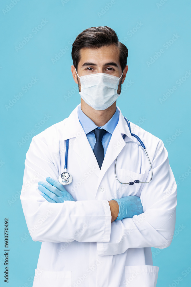 A male doctor in a white coat and a medical mask stands with his hands and looks at the camera on a blue isolated background, copy space, space for text A male doctor in a white coat and a medical mask stands with his hands and looks at the camera on a blue isolated background, copy space, space for text