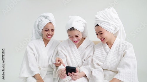 Group of young beautiful Asian woman in white bathrobe having fun and using smartphone sitting on bed in bedroom. Friends together concept