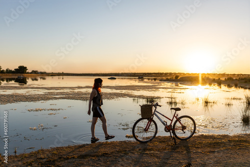 woman with bicycle on the lake at sunset in the middle of nature lake background and the sun
