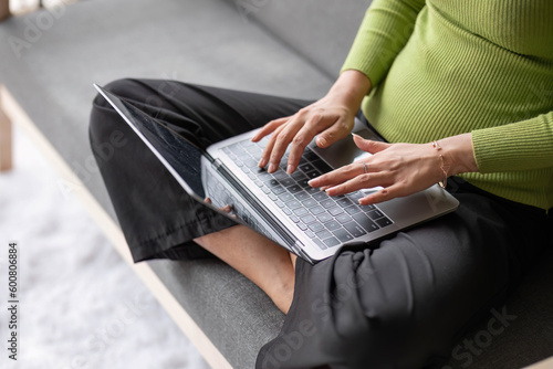 close-up image of a young Asian woman in casual clothes typing on keyboard, using her laptop computer on a sofa in her living room. laptop white screen mockup © chartchai