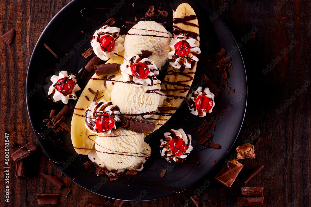 Vanilleeiskugeln mit Bananen und Kirschen auf Sahnetupfern mit Schokoroellchen auf schwarzem Teller und dunklem Holzfond als Topshot