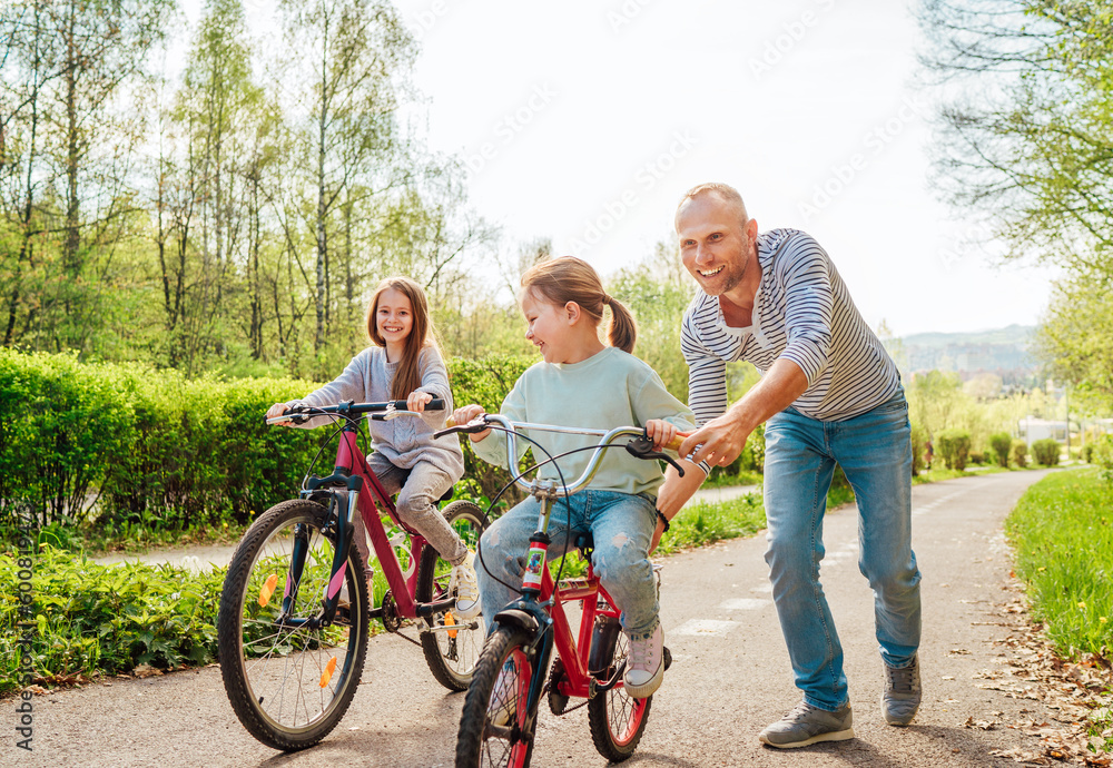 Smiling father with two daughters during outdoor walk. He teaching younger girl to ride a bicycle. They enjoy togetherness in the summer city park. Happy childhood concept image.