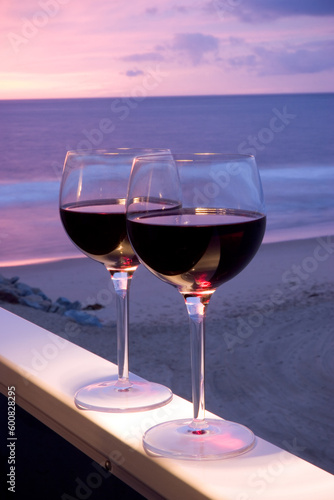Two glasses of red wine sitting on a ledge over looking the beach, ocean and beautiful sunset.