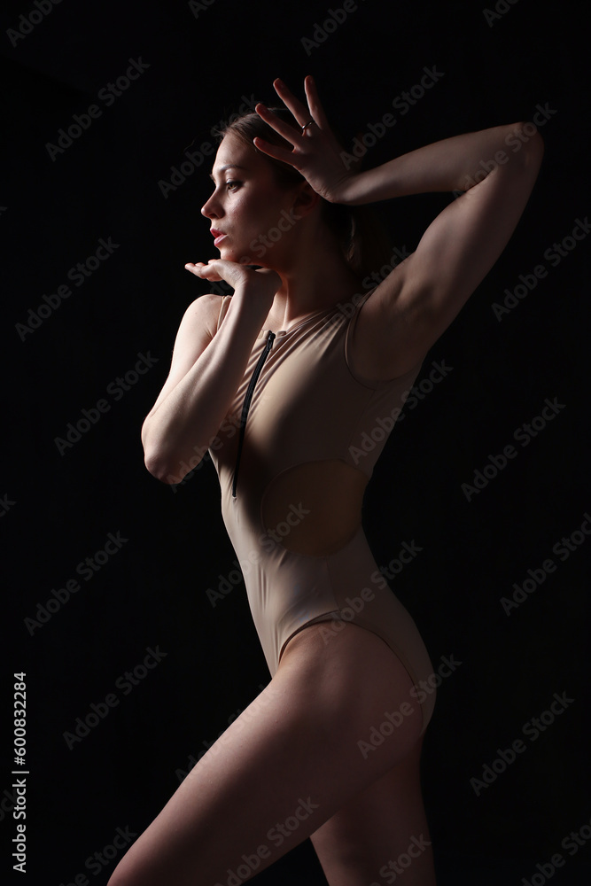 Obraz premium Ballerina in a beige bodysuit. Dark background. Sculpted beautiful female body. Pose of a gymnast.