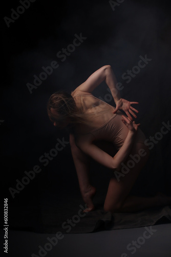 Ballerina in a beige bodysuit. Dark background. Sculpted beautiful female body. Pose of a gymnast.