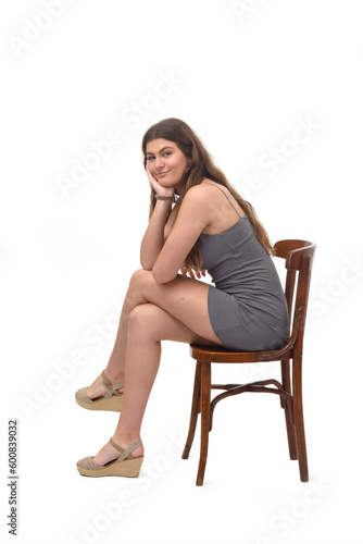 side view of young girl sitting on chair cross legged and hand on chin and looking at camera on white background