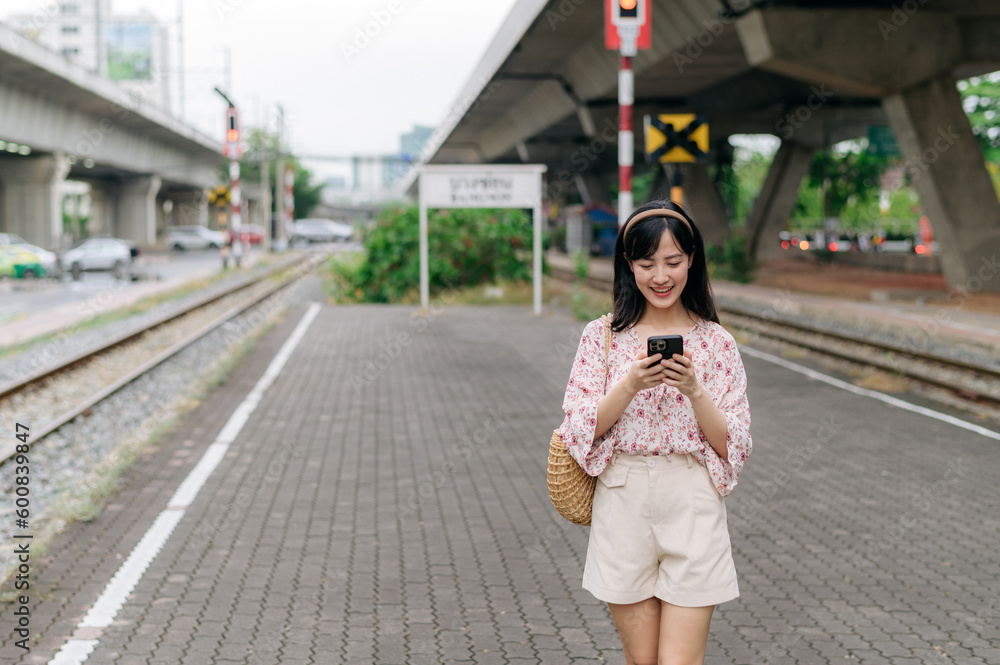 Asian young woman traveler with weaving basket using a mobile phone beside railway train station in Bangkok. Journey trip lifestyle, world travel explorer or Asia summer tourism concept. Asian young woman traveler with weaving basket using a mobile phone beside railway train station in Bangkok. Journey trip lifestyle, world travel explorer or Asia summer tourism concept.