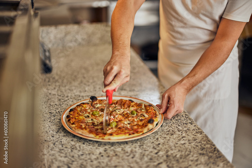 Close-up of a male worker, cutting a delicious pizza, using a pizza knife.