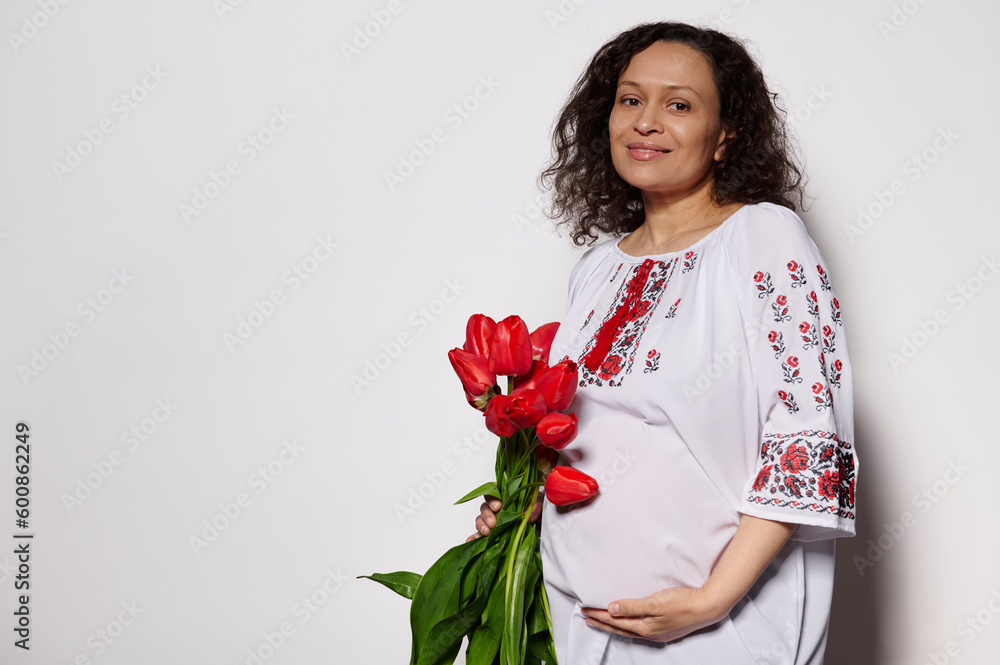 Happy delightful pregnant woman holding a bouquet of red tulips, caressing her belly, smiling, looking at camera on isolated white background, dressed in traditional ukrainian embroidered white dress Happy delightful pregnant woman holding a bouquet of red tulips, caressing her belly, smiling, looking at camera on isolated white background, dressed in traditional ukrainian embroidered white dress
