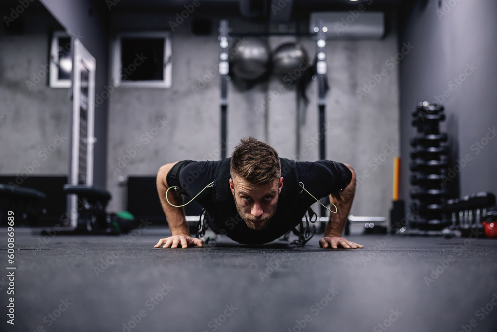 A male fitness coach in a special suit for EMS technology performing arm and chest exercise and push ups in a modern gym concept. Revolution in training, body rehabilitation - obrazy, fototapety, plakaty 