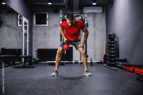 Hand training with sports equipment kettle bell. Full body portrait of a handsome and muscular man in the gym doing triceps exercises with kettle. Modern gym with a dark atmosphere, sports life