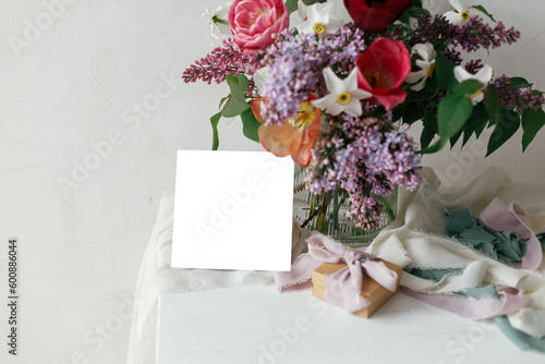 Greeting card mock up and lilac, tulips, daffodils flowers with present and pastel ribbons in modern rustic room. Happy Mothers day. Space for text. Stylish bouquet with gift box on wooden table