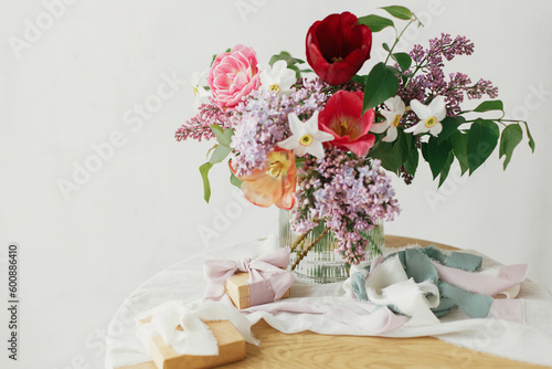 Stylish bouquet with gift box on rustic wooden table. Happy Mothers day. Spring lilac, tulips, daffodils arrangement with present on soft fabric in modern rustic room. Happy Womens day