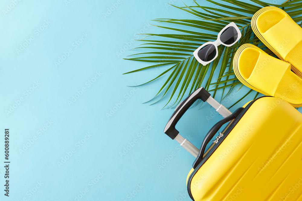 Summer travel concept. Top view flat lay of trendy yellow suitcase, summer flip flops, sunglasses and palm leaves on light blue background with empty space for text or advert