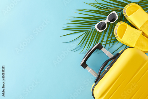 Summer travel concept. Top view flat lay of trendy yellow suitcase, summer flip flops, sunglasses and palm leaves on light blue background with empty space for text or advert