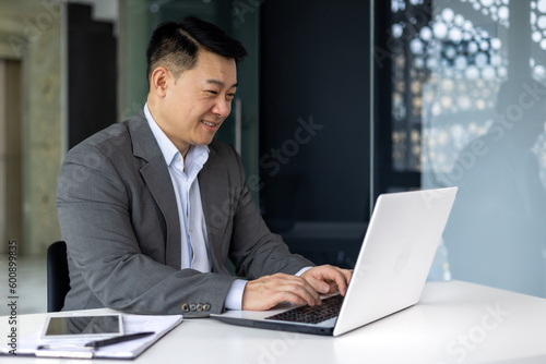 Successful mature asian working inside office using laptop, man typing on keyboard and smiling, businessman satisfied with work and achievement results, programmer at work.