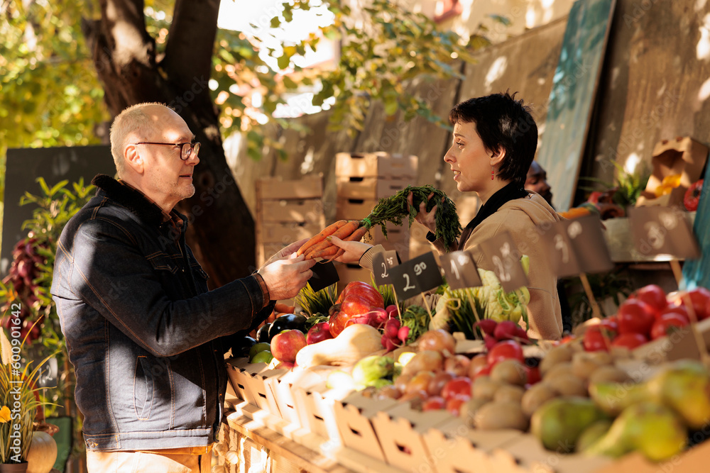 Woman seller showing organic natural carrots to elderly man at farmers market, locally grown. Small business owner standing behind fruit and vegetable stall selling healthy fresh produce to customer. Woman seller showing organic natural carrots to elderly man at farmers market, locally grown. Small business owner standing behind fruit and vegetable stall selling healthy fresh produce to customer.