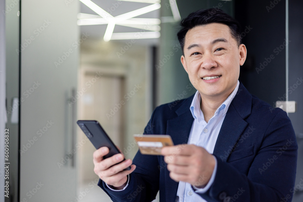 Closeup portrait of successful and satisfied asian man inside office, businessman smiling and looking at camera, mature man using money transfer app on phone, holding bank credit card. Closeup portrait of successful and satisfied asian man inside office, businessman smiling and looking at camera, mature man using money transfer app on phone, holding bank credit card.