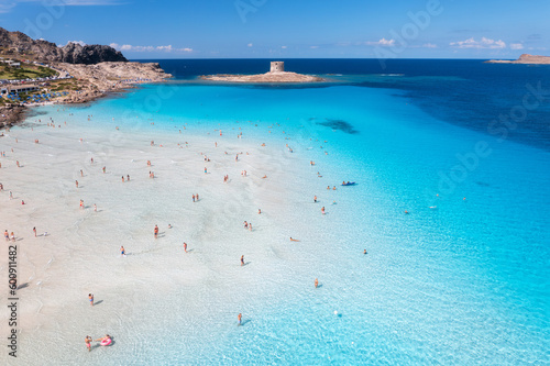Aerial view of famous La Pelosa beach at sunny summer day. Stintino, Sardinia island, Italy. Top view of sandy beach, swimming people, clear blue sea, old tower and sky with clouds. Tropical seascape