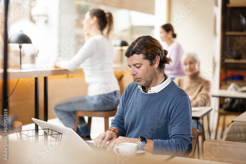 Positive man spending time in cozy coffeehouse with laptop and coffee