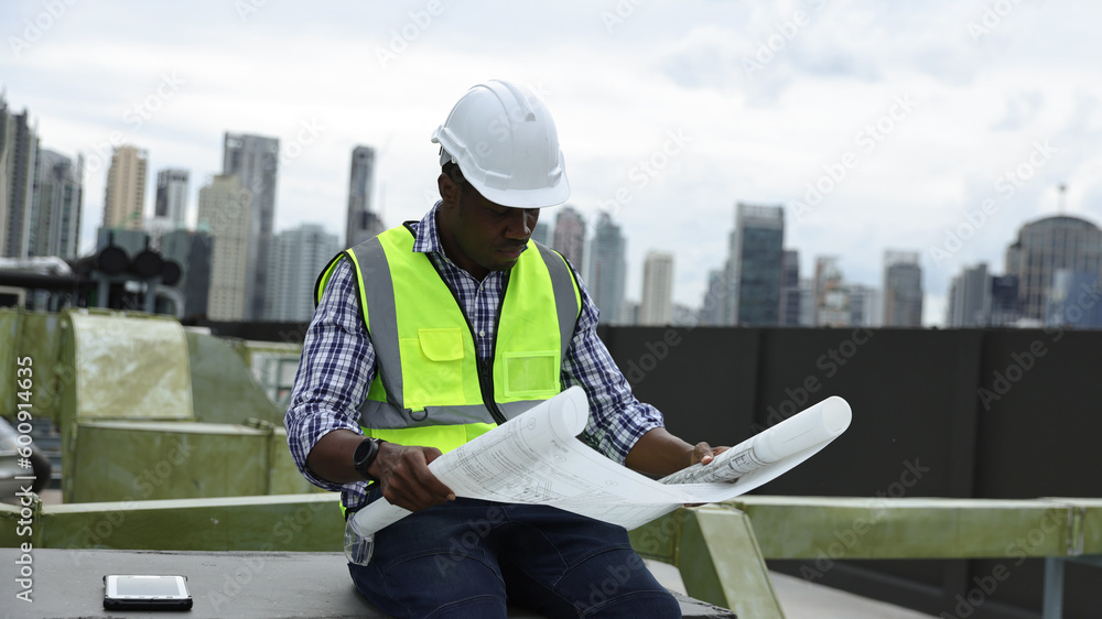 Black construction engineer holding blueprint working at construction site line. Mechanical engineer with safety vest and white safety helmet working with drawing at site line