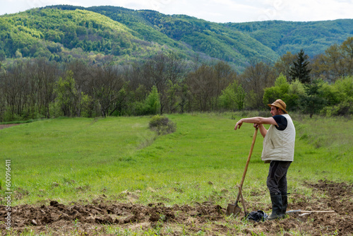 Farmer is standing leaning on his shovel and looking at the field he has plowed and prepared for planting vegetables. He is wearing a straw hat on his head. Agricultural concept