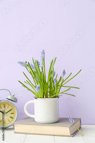 Cup with beautiful Muscari flowers  alarm clock and book on tiled table near lilac wall