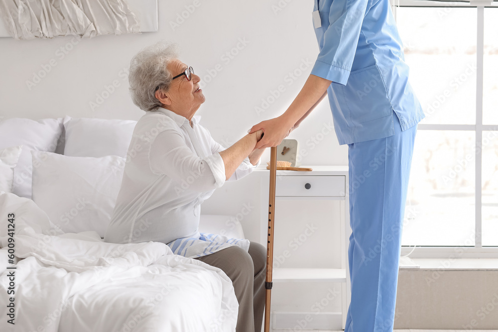 Obraz premium Young caregiver helping senior woman with stick to stand up in bedroom