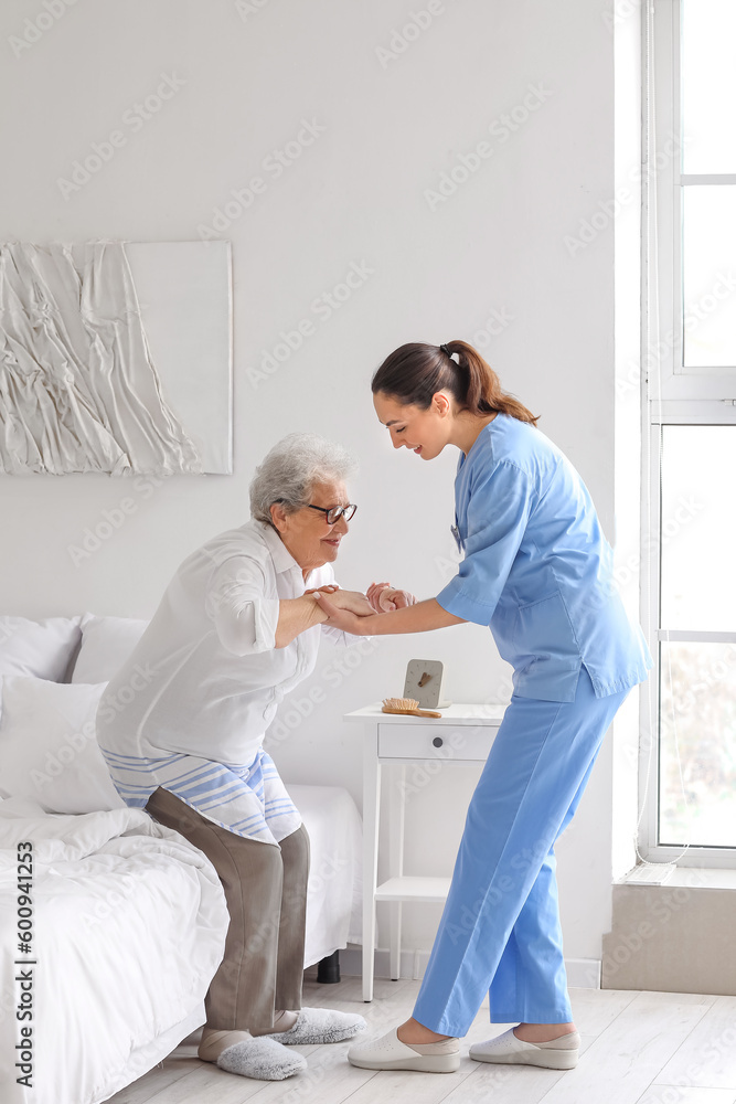 Young caregiver helping senior woman with stick to stand up in bedroom - obrazy, fototapety, plakaty 