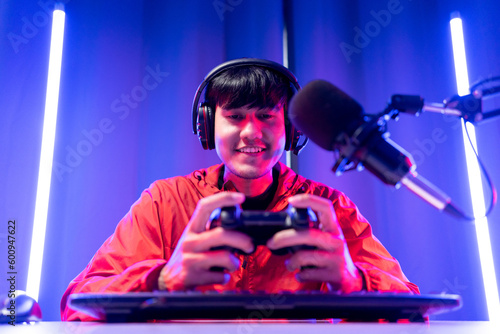 Professional gamer casting podcast walkthrough review. Young asian man sitting on chair with computer pc and microphone. Happy male Streamer wearing headphone playing game online in room neon light