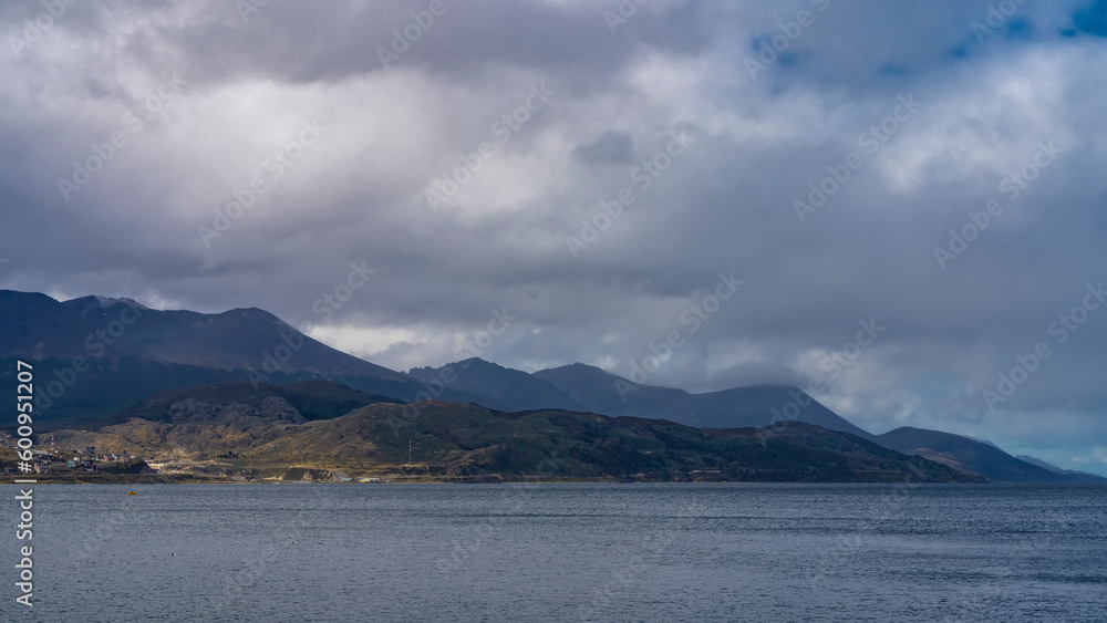 A picturesque mountain range against a cloudy sky. The blue water of the Beagle Canal in the foreground. City houses are visible on the shore. Argentina. Ushuaia. Tierra del Fuego archipelago.