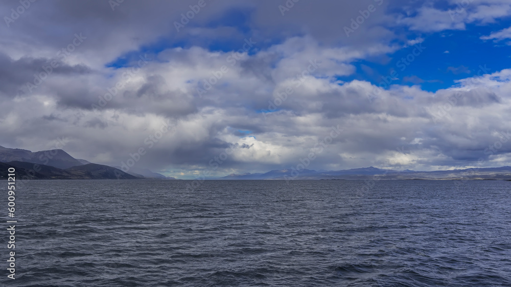 Ripples on the blue water of the Beagle Canal. In the distance, against the background of the azure sky and clouds - a mountain range. Argentina. Tierra del Fuego Archipelago. Ripples on the blue water of the Beagle Canal. In the distance, against the background of the azure sky and clouds - a mountain range. Argentina. Tierra del Fuego Archipelago.