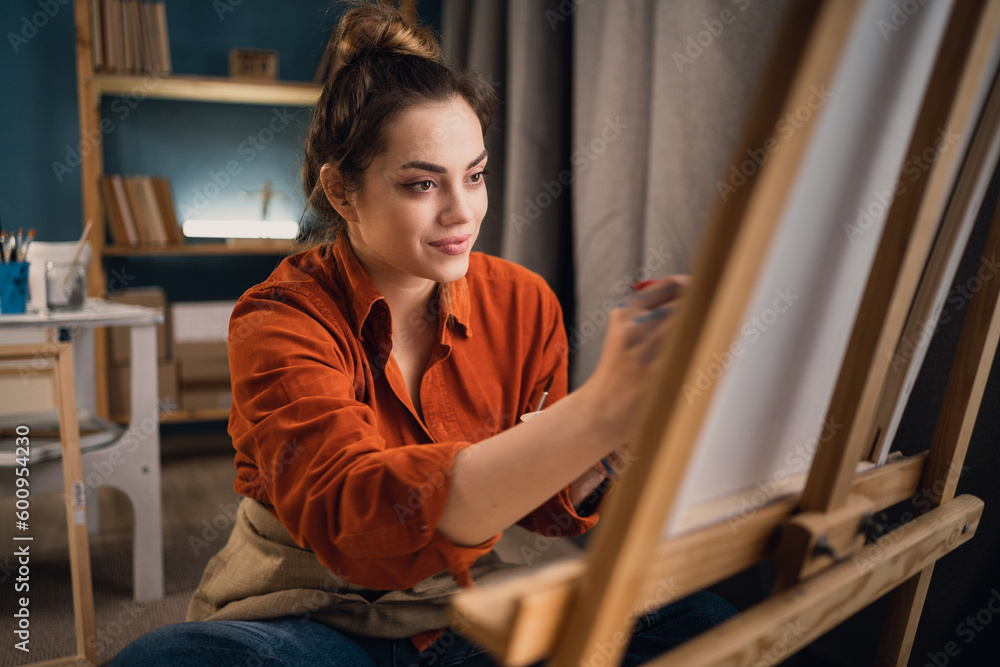 portrait of beautiful female artist, sits alone in bright modern workshop, paints picture on canvas with paints, looks absorbed, enjoys creative hobby for the soul. art class concept