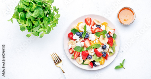 Sweet fruit berry salad with strawberries, blueberries, banana, soft cheese and mint leaves, white table background, top view