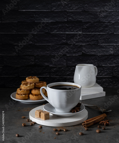 White chagka with black coffee stands on a light tray. Cinnamon tubes and coffee beans lie on the table. Biscuits on a saucer and milkman in the background. Dark brick background