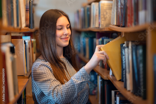 Young smiling girl standing near bookshelves in library taking books for reading, literature lesson. Science research, education, getting knowledge at university concept