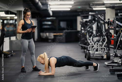 A happy female fitness instructor is holding tablet and looking at the sportswoman who is doing planks in a gym.