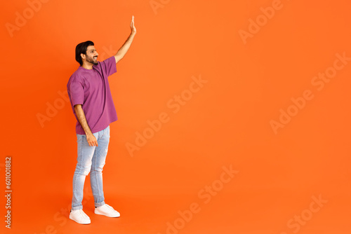 Cheerful eastern guy waving at copy space, orange background