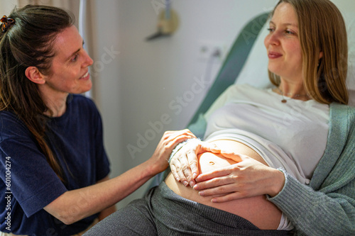 Lovely young lesbian couple planning the future of their baby, woman smiling with hands on expecting mother's baby bump while feeling baby movement, assisted fertilization and ivf photo