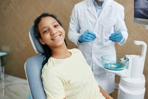 High angle portrait of smiling black girl sitting in dental chair and looking at camera during checkup