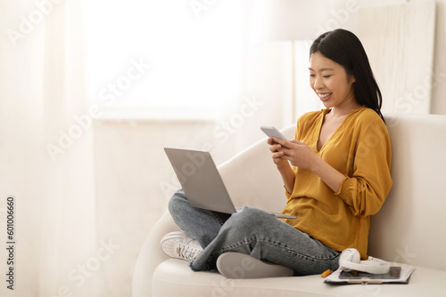Happy young asian woman sitting on couch, using gadgets © Prostock-studio