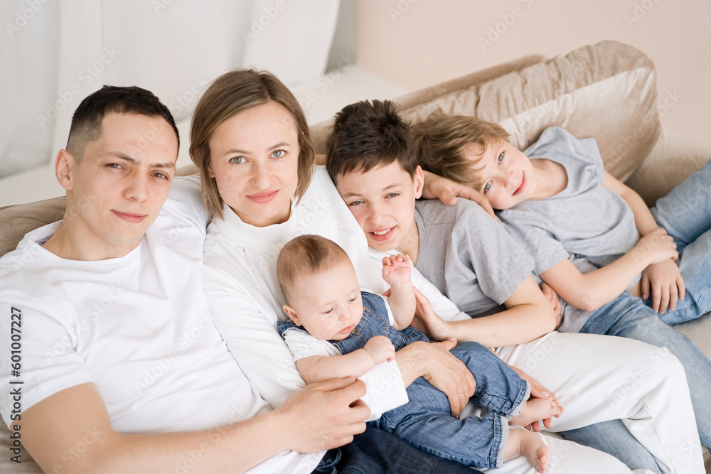 Portrait of smiling mother and father with
three sons sitting on the couch and looking at the camera, happy parents hugging children, big family posing for a photo at home Portrait of smiling mother and father with
three sons sitting on the couch and looking at the camera, happy parents hugging children, big family posing for a photo at home