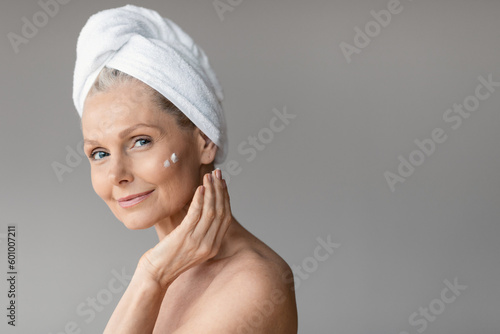 Morning beauty treatments for aged skin. Happy senior woman in towel applying cream under eyes after shower, copy space