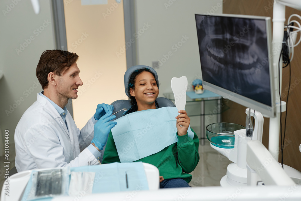 Fototapeta premium Portrait of black teenage girl at dental checkup in clinic looking n mirror and smiling