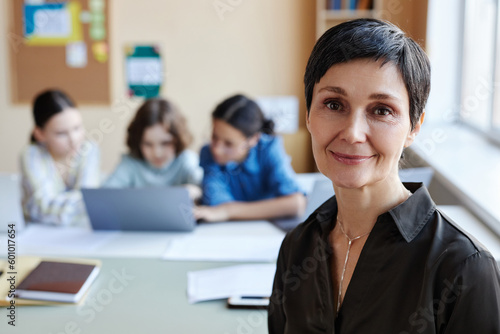 Portrait of professional teacher smiling at camera while sitting at lesson with children in background