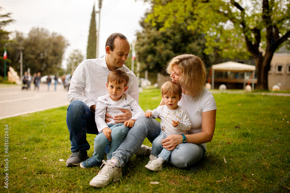 Parents and their small toddler kids son and daughter sitting on the green grass in the park and hugging each other; happy family concept Parents and their small toddler kids son and daughter sitting on the green grass in the park and hugging each other; happy family concept