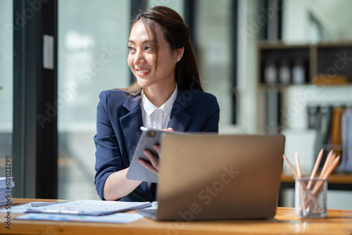 Asian businesswoman in a suit sitting holding iPad or tablet in contact See details and discuss business information happily in the office.