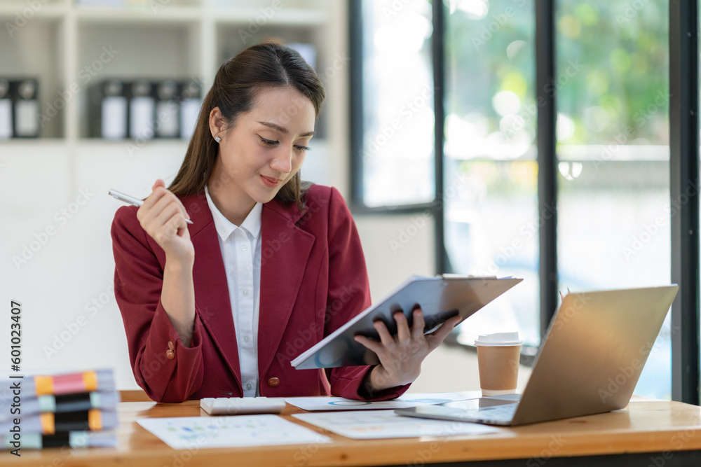 Beautiful attractive Asian businesswoman holding a clipboard of documents sitting analytically analyzing various marketing management information the finances were at her desk in the office at ease. Beautiful attractive Asian businesswoman holding a clipboard of documents sitting analytically analyzing various marketing management information the finances were at her desk in the office at ease.