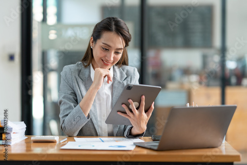 Asian businesswoman in a suit sitting holding iPad or tablet in contact See details and discuss business information happily in the office.